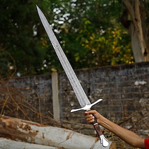 May include: A long, silver sword with a patterned blade and a cross-shaped hilt. The handle is brown with a silver pommel. The sword is held up against a backdrop of trees and a brick wall.