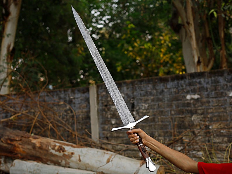 May include: A long, silver sword with a patterned blade and a cross-shaped hilt. The handle is brown with a silver pommel. The sword is held up against a backdrop of trees and a brick wall.