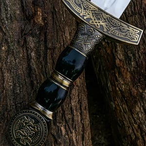 May include: A decorative sword with a silver blade and an ornate handle. The handle has a dark green grip with gold accents and a detailed, bronze-coloured guard and pommel. The sword is set against a textured, brown tree trunk.
