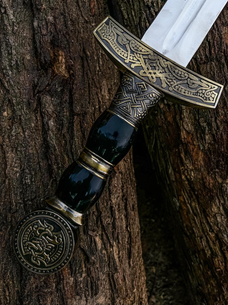 May include: A decorative sword with a silver blade and an ornate handle. The handle has a dark green grip with gold accents and a detailed, bronze-coloured guard and pommel. The sword is set against a textured, brown tree trunk.