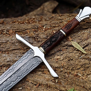 May include: A detailed close-up of a sword with a patterned blade and a brown wooden handle. The handle features intricate black designs and is accented with silver and gold-colored metal. The sword rests on a textured wooden surface.