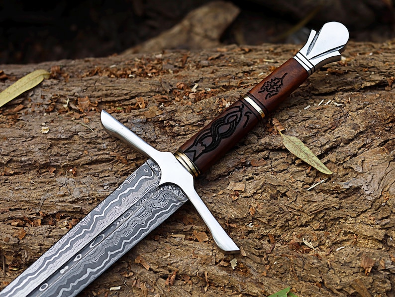 May include: A detailed close-up of a sword with a patterned blade and a brown wooden handle. The handle features intricate black designs and is accented with silver and gold-colored metal. The sword rests on a textured wooden surface.