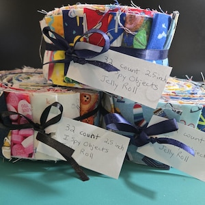 May include: Three fabric jelly rolls featuring assorted colorful patterns, each tied with a dark blue ribbon and a white paper tag. The tags are handwritten with the text 32 count 2.5 inch I Spy Objects Jelly Roll. The fabric strips are rolled tightly.