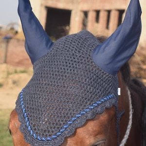 Può includere: Un cappuccio per cavallo all'uncinetto grigio scuro con paraorecchie blu navy e un bordo in corda blu. Il cappuccio è progettato per proteggere le orecchie del cavallo da insetti e rumori. La testa del cavallo è visibile.