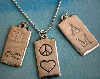 May include: A silver chain necklace with three silver rectangular pendants. The pendants have different designs: one has the letter "H" and an infinity symbol, another has a peace sign and a heart, and the third has the letters "AM".