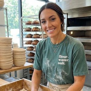 May include: A baker in a green t-shirt with the words "flour. water. salt." kneads dough in a wooden trough. Stacks of round, woven baskets and loaves of bread are visible in the background, suggesting a bakery setting.