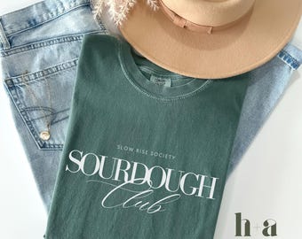 May include: A green t-shirt with the text "SOURDOUGH Club" in white lettering. The text "Slow Rise Society" is printed above the main text. The text "h+a HARVEST + ANCHOR CO. SINCE 2014" is printed in white on a black rectangle in the bottom right corner. The text "BLUE SPRUCE" is printed in white below the rectangle.