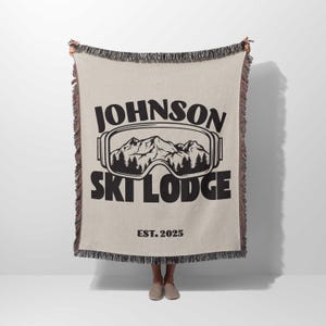 May include: A woven throw blanket with a black and white design featuring the text "Johnson Ski Lodge Est. 2025" and a pair of ski goggles with a mountain scene inside.