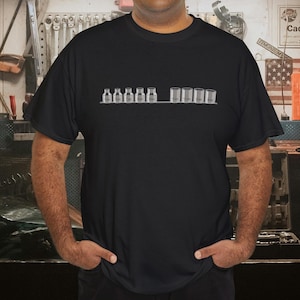 Könnte beinhalten: Schwarzes T-Shirt mit einer minimalistischen Grafik von Glasflaschen und zylindrischen Behältern. Das Design ist horizontal angeordnet. Das T-Shirt ist aus einem dunklen Stoff und wird getragen.