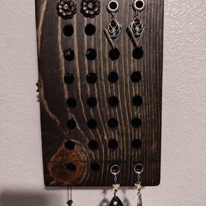 May include: A dark wood earring holder with multiple holes for hanging earrings. The holder has a few earrings hanging from it, including a pair of silver earrings with a black and white design, a pair of silver earrings with a star design, and a pair of silver earrings with a blue and white design.