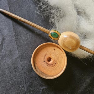 May include: A wooden drop spindle featuring a long, tapered shaft with a smooth, light-colored wooden whorl and a smaller, multi-colored accent bead. A separate, circular wooden bowl with a central indentation sits below the spindle.