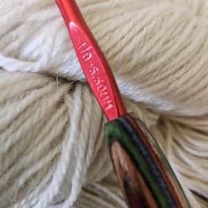 May include: A red crochet hook with a wooden handle, resting against a ball of cream-colored yarn. The hook is marked with the size 1/9 - 5.50mm. The handle features a striped pattern of green, brown, and red.