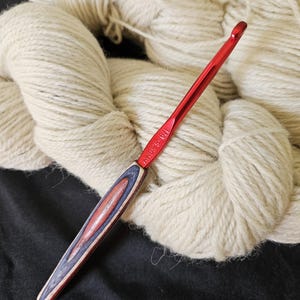 May include: A red crochet hook with a wooden handle featuring red, blue, and natural wood tones. The hook is positioned over a background of off-white yarn, suggesting a crafting theme.