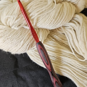 May include: A red crochet hook with a wooden handle featuring a red and black grain pattern. The hook is positioned in front of a large ball of off-white yarn, suggesting a crafting theme.