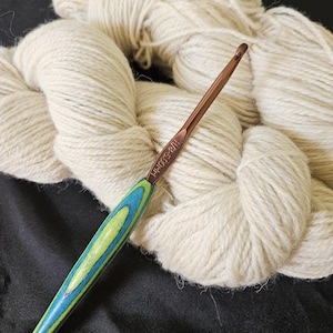 May include: A crochet hook with a copper-colored metal head and a handle with blue and green wood grain. The hook is next to a large skein of off-white yarn, suggesting a crafting theme. The hook is labeled H/8-5.00MM.