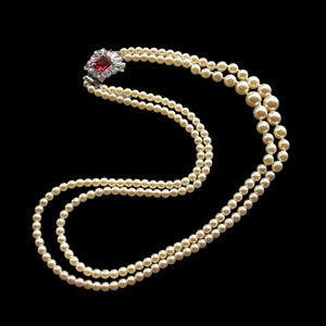 May include: A double-strand pearl necklace with a decorative clasp. The clasp features a central red gemstone surrounded by smaller clear stones. The pearls are off-white and the background is black.