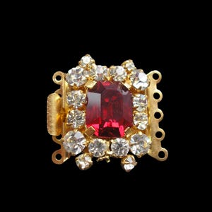 Peut inclure: Fermoir doré avec un strass rouge au centre entouré de strass transparents.