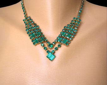 May include: Emerald green rhinestone necklace with a gold-tone setting. The necklace features a V-shaped design with rectangular and round stones. A large square stone hangs at the center. The necklace is displayed on a mannequin.
