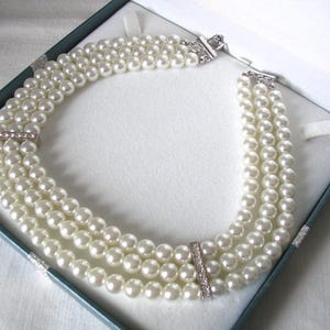 May include: A three-strand pearl necklace with a silver clasp and rhinestone detailing, displayed in a blue jewellery box. The necklace is composed of creamy white pearls, offering a timeless and sophisticated aesthetic.