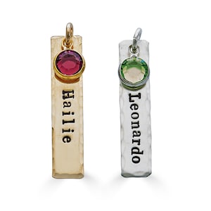 May include: Two personalized bar pendants. One is gold-toned with a red gemstone and the name "Hailie". The other is silver-toned with a green gemstone and the name "Leonardo". Both pendants have a hammered texture and a loop at the top.