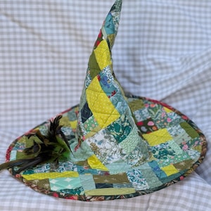 May include: A quilted witch hat constructed from a patchwork of various fabric squares in shades of green, yellow, and blue. The hat features a tall, pointed crown and a wide brim, accented with a cluster of dark feathers on the left side.