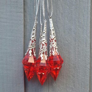 May include: Three red, faceted crystal pendants with ornate silver-colored metal caps and silver cords hang against a gray wooden background. The pendants are teardrop-shaped and reflect light.