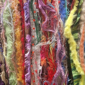 May include: A close-up of a colourful assortment of fabric scraps, ribbons, and yarn. The materials are arranged vertically and feature a variety of textures and colours, including green, blue, red, purple, and yellow.