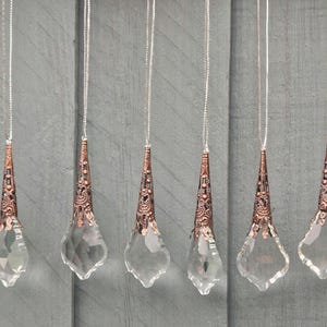 May include: Six clear crystal pendants with copper-coloured filigree caps and silver chains hang against a grey wooden background. The pendants have a teardrop shape with a pointed top and a rounded bottom.