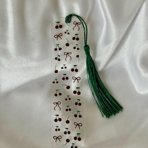 May include: A rectangular clear acrylic bookmark featuring a repeating pattern of small dark red cherries and thin dark red bows. A dark green thread tassel is attached through a hole at the top of the bookmark, providing a decorative accent.