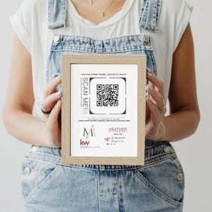May include: A framed print with a QR code and the text "SCAN ME". The print also includes contact information for a realtor. The frame is light brown, and the print is held by a person wearing denim overalls.