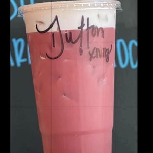 May include: A clear plastic disposable cup filled with a pink iced beverage topped with a layer of white foam. The cup features a clear plastic lid and has the name Dutton and the letters xnrg written in black marker on the side of the cup.