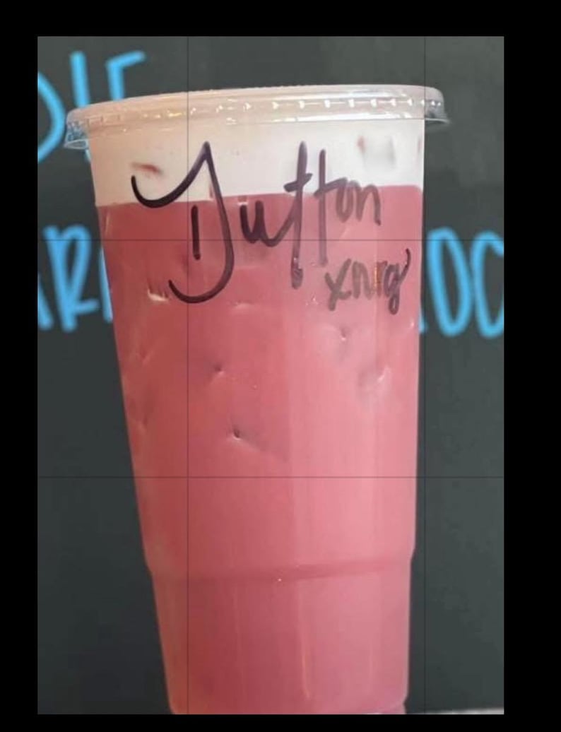 May include: A clear plastic disposable cup filled with a pink iced beverage topped with a layer of white foam. The cup features a clear plastic lid and has the name Dutton and the letters xnrg written in black marker on the side of the cup.