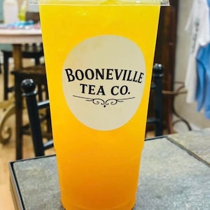 May include: A clear plastic disposable cup filled with a bright yellow iced beverage. The cup features a circular white label on the front with the black text BOONEVILLE TEA CO. printed above a decorative horizontal line and swirl design.