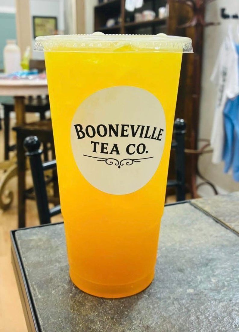 May include: A clear plastic disposable cup filled with a bright yellow iced beverage. The cup features a circular white label on the front with the black text BOONEVILLE TEA CO. printed above a decorative horizontal line and swirl design.