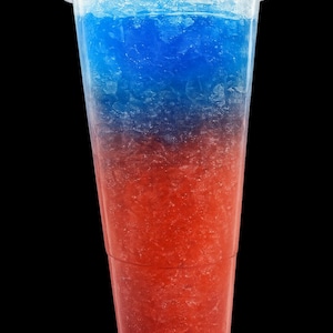 May include: A clear plastic disposable cup filled with a layered frozen slushy beverage. The drink features a distinct horizontal color gradient, transitioning from a vibrant blue at the top to a bright red at the bottom, with crushed ice texture.
