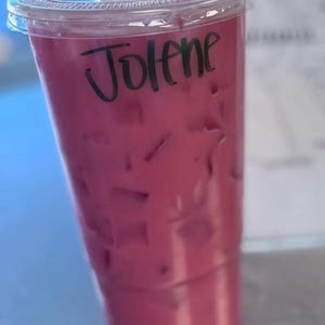 May include: A clear plastic disposable cup filled with a vibrant magenta iced beverage. The cup is topped with a matching clear plastic lid and features the name Jolene written in black marker on the side of the cup above the liquid line.