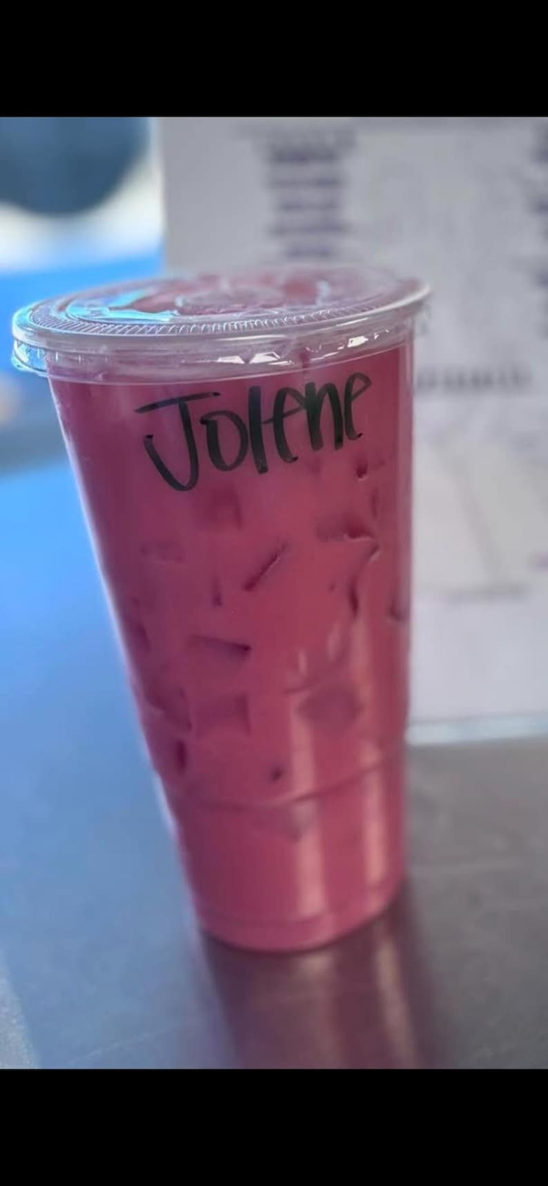 May include: A clear plastic disposable cup filled with a vibrant magenta iced beverage. The cup is topped with a matching clear plastic lid and features the name Jolene written in black marker on the side of the cup above the liquid line.