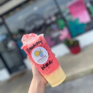 May include: A clear plastic cup filled with a layered pink and yellow beverage, topped with whipped cream and pink sprinkles. The cup features the handwritten text Strawberry blonde and a circular logo for Sunshine and Loaded Teas.