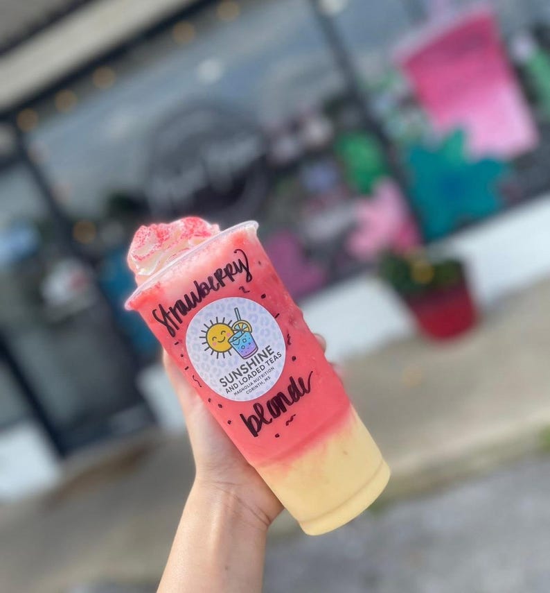 May include: A clear plastic cup filled with a layered pink and yellow beverage, topped with whipped cream and pink sprinkles. The cup features the handwritten text Strawberry blonde and a circular logo for Sunshine and Loaded Teas.
