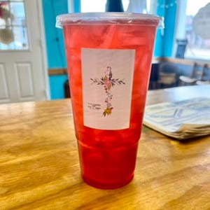 May include: A clear plastic disposable cup filled with a bright red iced beverage and ice cubes. A rectangular white label is attached to the front, featuring a floral cross design composed of small colorful flowers and green leaves.