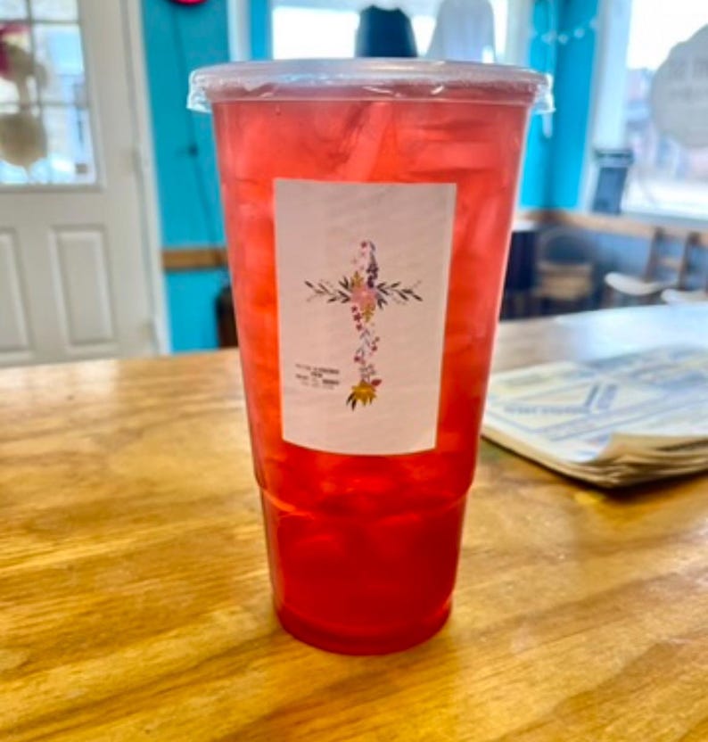 May include: A clear plastic disposable cup filled with a bright red iced beverage and ice cubes. A rectangular white label is attached to the front, featuring a floral cross design composed of small colorful flowers and green leaves.