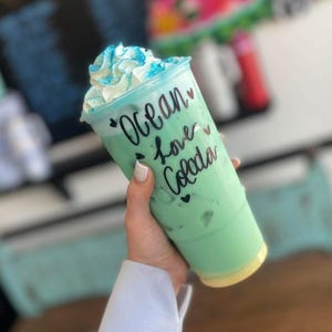 May include: A clear plastic tumbler filled with a light green beverage and ice, topped with white whipped cream and blue sprinkles. The cup features black cursive text that reads Ocean love Colada, accented with small black heart symbols.
