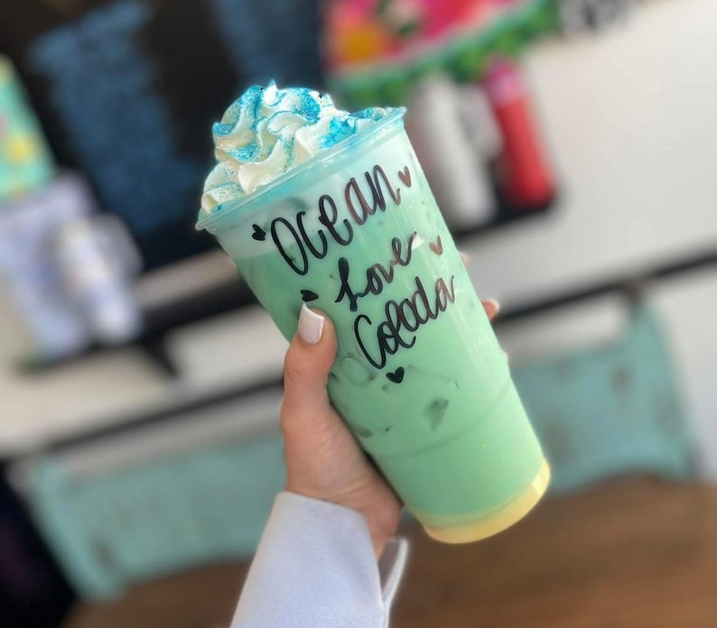 May include: A clear plastic tumbler filled with a light green beverage and ice, topped with white whipped cream and blue sprinkles. The cup features black cursive text that reads Ocean love Colada, accented with small black heart symbols.