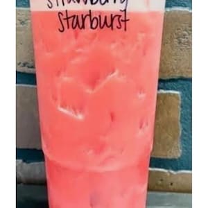 May include: A clear plastic cup filled with a pink iced beverage and ice cubes. The top portion of the cup features a layer of white foam. The text "Strawberry Starburst" is written in black marker on the upper section of the clear plastic cup.