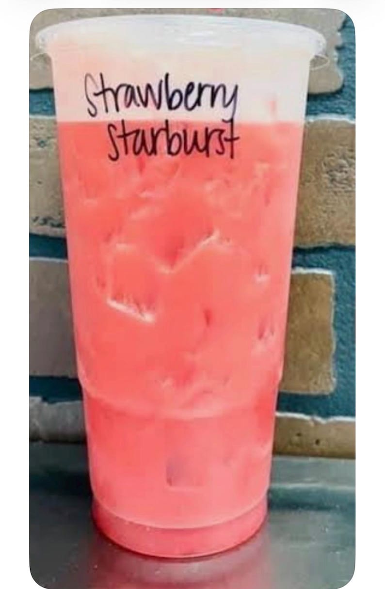 May include: A clear plastic cup filled with a pink iced beverage and ice cubes. The top portion of the cup features a layer of white foam. The text "Strawberry Starburst" is written in black marker on the upper section of the clear plastic cup.