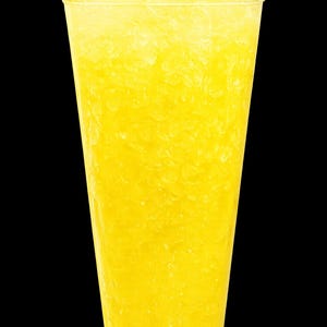 May include: A clear plastic cup filled with a bright yellow, icy beverage. The drink is packed with crushed ice, creating a textured, slushy appearance that fills the container from the bottom to the top rim against a solid black background.