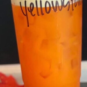 May include: A clear plastic disposable cup filled with an orange liquid and ice cubes. The word Yellowstone is written in black marker on the side of the cup. The cup has a clear plastic lid and a layer of white foam at the top of the liquid.