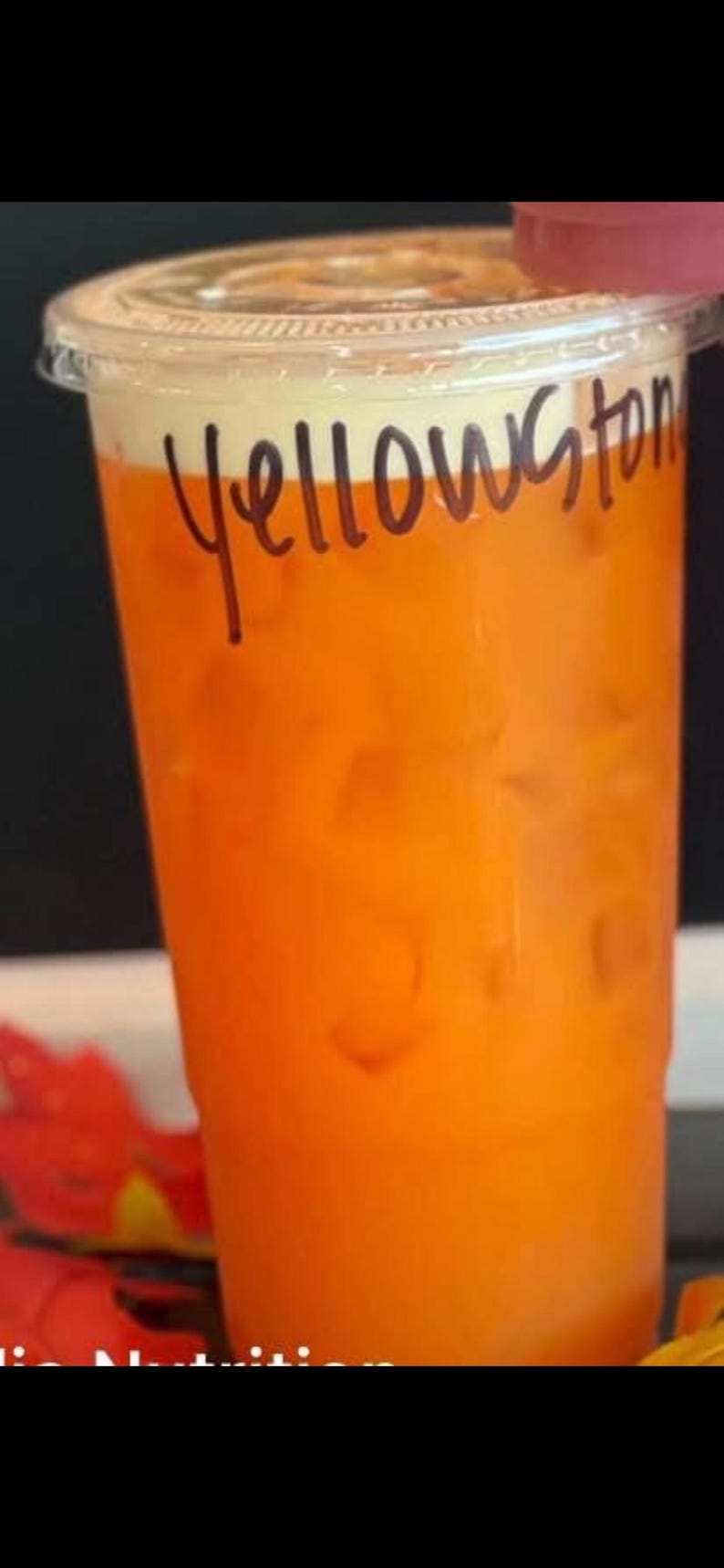 May include: A clear plastic disposable cup filled with an orange liquid and ice cubes. The word Yellowstone is written in black marker on the side of the cup. The cup has a clear plastic lid and a layer of white foam at the top of the liquid.