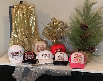 May include: A collection of trucker hats with various designs and colors. The hats feature text and graphics, including Christmas themes, a Friends theme, and other decorative elements. The hats are displayed on a black surface.