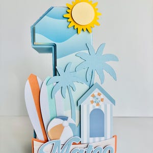 May include: A beach-themed number one cake topper with a yellow sun, palm trees, a surfboard, and a beach ball. The name "Mateo" is displayed in cursive script. The topper is made of layered paper in shades of blue, orange, and white.